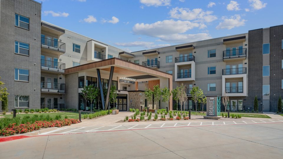 the exterior of an apartment complex with a driveway and landscaping at The BroadVue